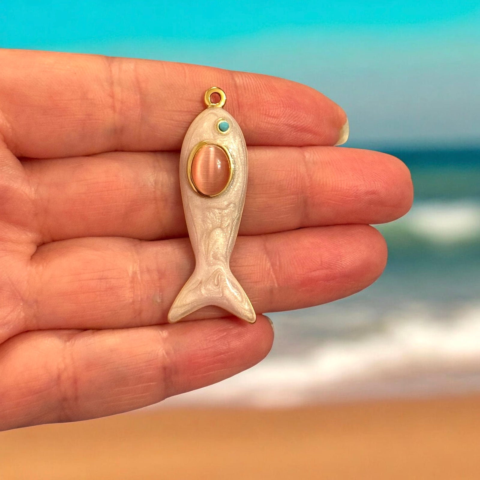 Stainless Steel 24K PVD Gold Plated Blush Pink Fish Pendant held in hand by beach with ocean background.Stainless Steel 24K PVD Gold Plated Blush Pink Fish Pendant